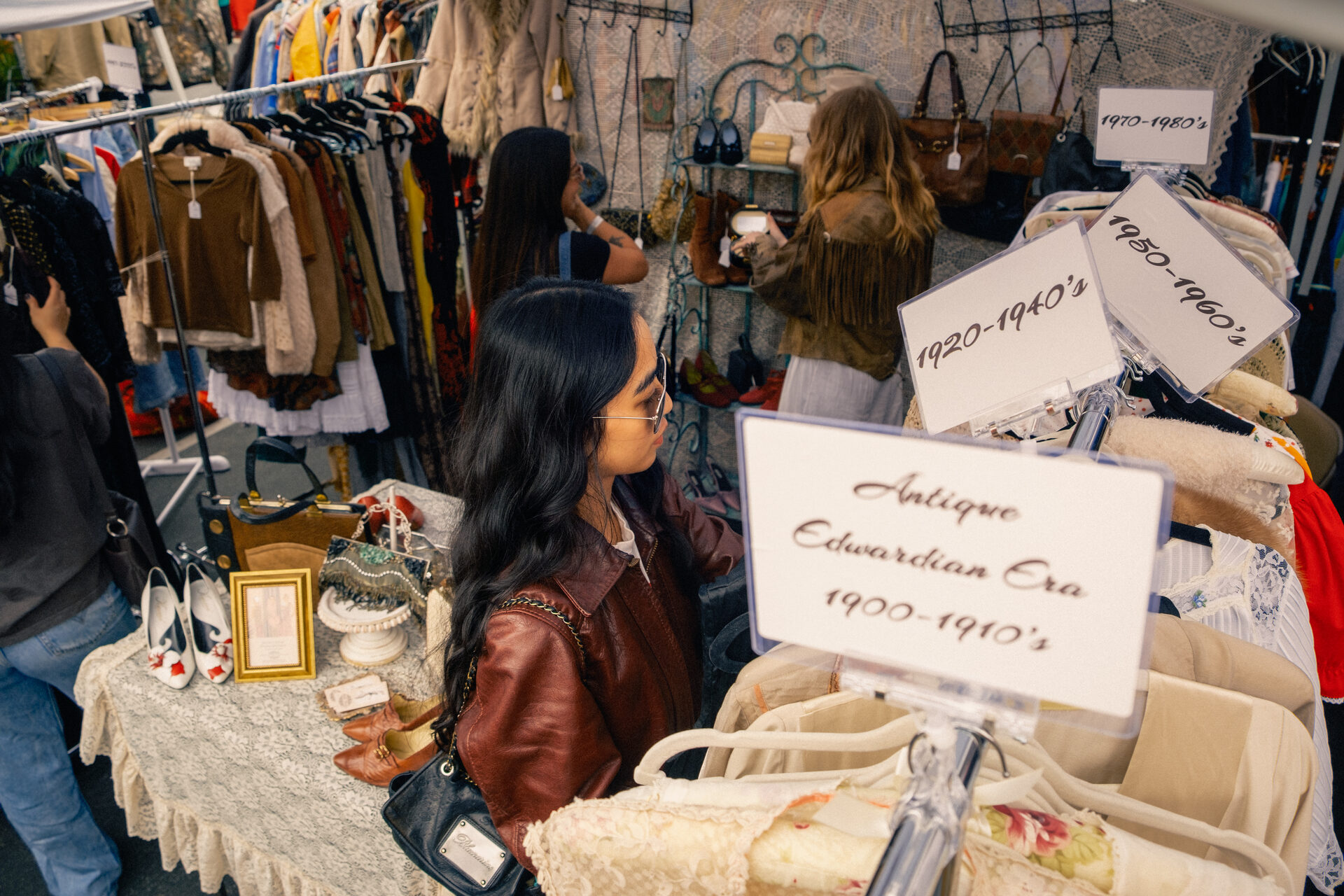 A shopper in a leather jacket and sunglasses browses an Antique Edwardian Era 1900-1910's clothing rack inside a curated vintage booth at the Los Feliz Flea, with hand-lettered decade signs (1920-1940's, 1950-1960's, 1970-1980's) marking the racks